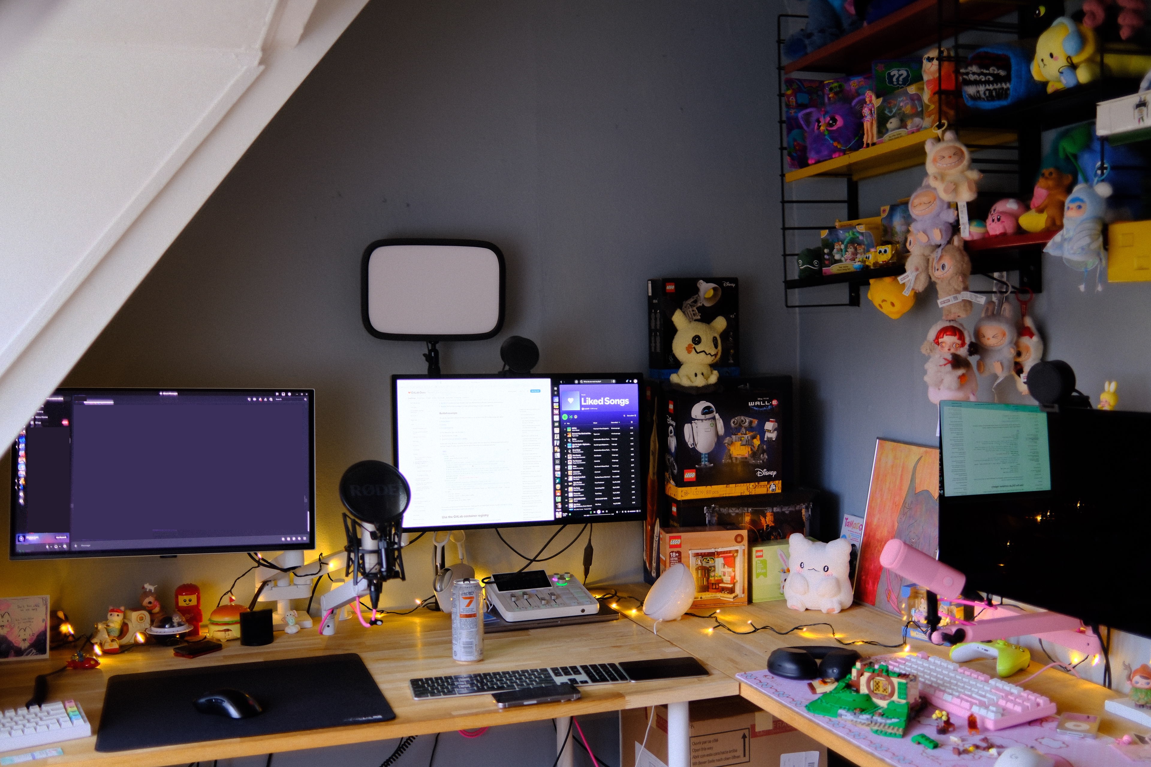 A home desk setup tucked under a sloped ceiling. Two monitors sit side by side — the left one dark, the right one displaying a document next to a Spotify 'Liked Songs' playlist — with a Rode podcast microphone on a boom arm and a rectangular key light mounted above. A Lego WALL-E set is stacked on boxes to the right of the center monitor. The right side of the desk holds a pink mechanical keyboard, black headphones, and a glowing cat-shaped lamp, with warm fairy lights draped across the wooden desktop. A wire wall shelf in the upper right is packed with colorful plush toys and Sonny Angel figures.